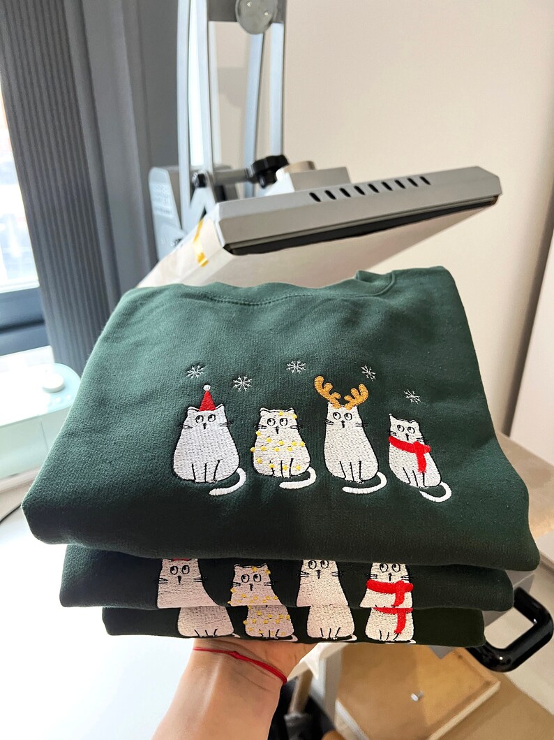 Peut inclure: Des sweat-shirts vert fonc&eacute; avec des chats blancs brod&eacute;s portant des accessoires de f&ecirc;te. Les chats sont orn&eacute;s d'un bonnet de P&egrave;re No&euml;l, de guirlandes lumineuses, de bois de renne et d'une &eacute;charpe. Les sweat-shirts sont empil&eacute;s, mettant en valeur le motif.