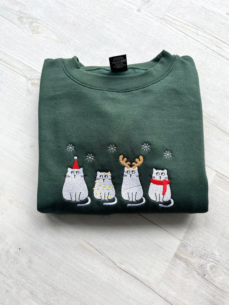 Peut inclure: Un sweat-shirt vert fonc&eacute; pli&eacute;, orn&eacute; d'une broderie de quatre chats blancs avec des accessoires festifs. Les chats sont orn&eacute;s d'un bonnet de P&egrave;re No&euml;l, de guirlandes lumineuses, de bois de renne et d'une &eacute;charpe rouge, avec des flocons de neige brod&eacute;s.