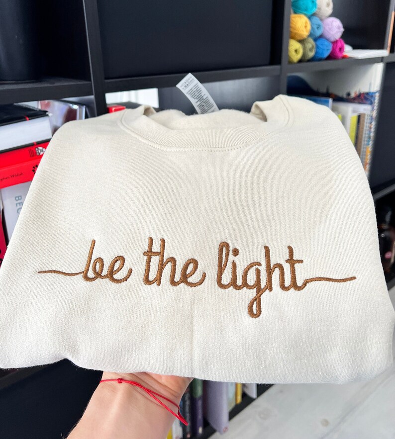 Op de afbeelding: Cr&egrave;mekleurige sweatshirt met de geborduurde tekst "be the light" in bruin. De opgevouwen sweatshirt toont de tekst duidelijk. De stof ziet er zacht en comfortabel uit, ideaal voor dagelijks gebruik.