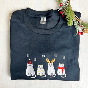 Peut inclure: T-shirt noir avec un motif brod&eacute; de quatre chats de dessin anim&eacute; avec des accessoires de f&ecirc;te et des flocons de neige. Les chats portent un bonnet de P&egrave;re No&euml;l, des lumi&egrave;res, des bois de renne et une &eacute;charpe. Th&egrave;me des f&ecirc;tes.