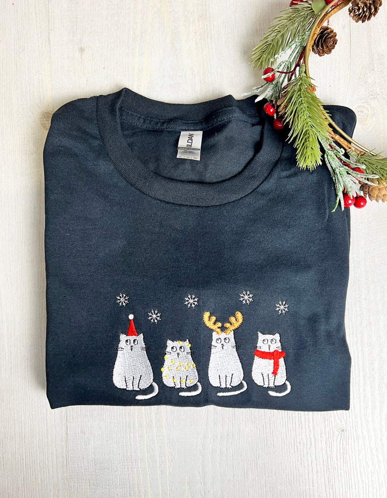 Peut inclure: T-shirt noir avec un motif brod&eacute; de quatre chats de dessin anim&eacute; avec des accessoires de f&ecirc;te et des flocons de neige. Les chats portent un bonnet de P&egrave;re No&euml;l, des lumi&egrave;res, des bois de renne et une &eacute;charpe. Th&egrave;me des f&ecirc;tes.