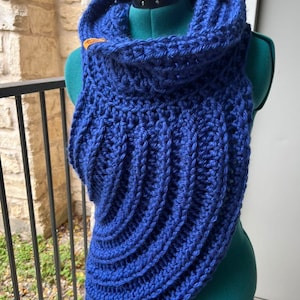 May include: A handmade, blue crochet cowl neck warmer. The cowl is displayed on a mannequin and features a ribbed design. A small, brown leather tag is attached near the neck. The cowl is a cozy accessory for cold weather.