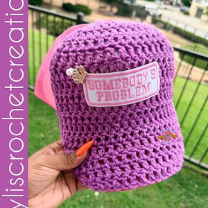 Op de afbeelding: Een paarse gehaakte honkbalpet met een roze mesh achterkant en een rechthoekige patch met de tekst SOMEBODY'S PROBLEM in roze letters. Een kleine goudkleurige kroonbedel met een witte parel is aan de linkerkant van de patch bevestigd.