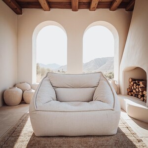 Puede incluir: Un sill&oacute;n bajo y mullido tapizado en tela de color beige claro con un fino patr&oacute;n de rayas verticales. La silla presenta una silueta redondeada de bordes suaves, con un asiento profundo, reposabrazos integrados y un respaldo acolchado.