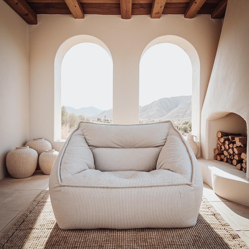 Puede incluir: Un sill&oacute;n bajo y mullido tapizado en tela de color beige claro con un fino patr&oacute;n de rayas verticales. La silla presenta una silueta redondeada de bordes suaves, con un asiento profundo, reposabrazos integrados y un respaldo acolchado.