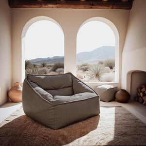 Puede incluir: Un sill&oacute;n bajo de color topo con un acabado de tela texturizada y acanalada. El sill&oacute;n presenta una silueta estructurada y cuadrada con un asiento profundo y un respaldo ligeramente reclinado, colocado sobre una alfombra tejida en una estancia.
