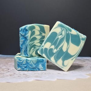 Puede incluir: Cuatro barras de jabón hechas a mano con un diseño de remolino azul y blanco. Las barras de jabón están apiladas y dispuestas sobre una superficie arenosa con un fondo oscuro. El jabón tiene una apariencia veteada.