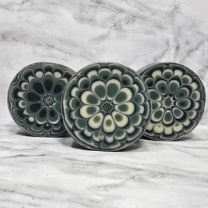 Puede incluir: Tres jabones redondos con un diseño floral. Los jabones presentan un patrón de pétalos verdes oscuros, verdes claros y blancos. Los jabones están dispuestos sobre una superficie de mármol.
