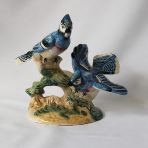 May include: A ceramic ornament depicting two blue jays on a branch. The birds are predominantly blue with black and white markings and red details. The branch is painted green and brown, and the base is a light tan colour.