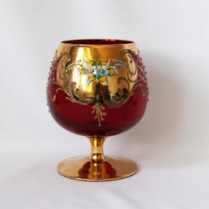 May include: A red and gold brandy snifter with floral and scrollwork designs. The glass features a gold rim and base, with a red bowl. The bowl is decorated with gold filigree and painted flowers. The snifter is likely made of glass.