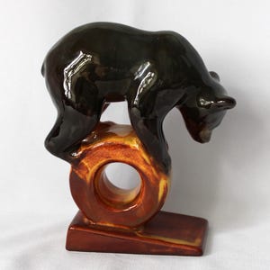 May include: A ceramic figurine of a bear in a climbing pose. The bear is dark brown with a glossy finish. It is perched on a ring-shaped structure with a base, featuring shades of brown and yellow.