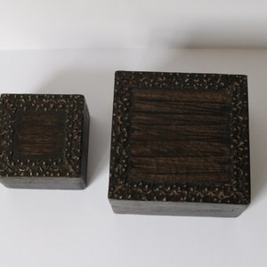 May include: Two wooden boxes with intricate carved designs around the edges. The boxes are dark brown and have a natural wood grain. The smaller box is on the left and the larger box is on the right.