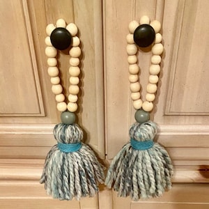 May include: Two wooden bead tassels with gray and blue yarn pom-poms. The tassels are attached to cabinet doors.