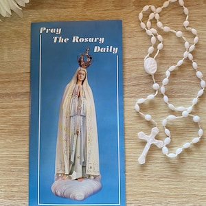 May include: A blue pamphlet with the text "Pray The Rosary Daily" and a picture of the Virgin Mary in a white robe with a crown on her head. A white rosary with a white cross is on a wooden surface.