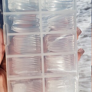 May include: A clear plastic storage box with multiple compartments filled with clear nail tips. The box has a handle on the top.