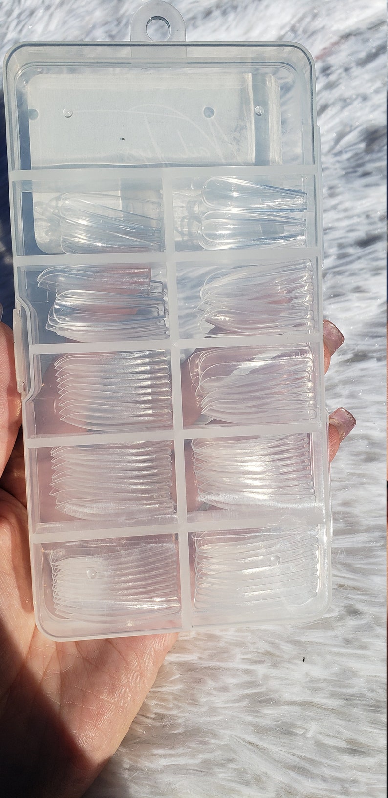 May include: A clear plastic storage box with multiple compartments filled with clear nail tips. The box has a handle on the top.