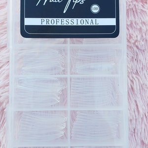 May include: A clear plastic box with a black label that says "Nail Tips Professional (100)". The box contains 6 compartments filled with clear plastic nail tips.