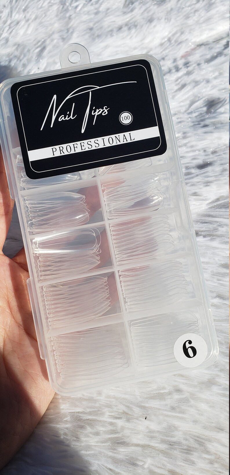 May include: A clear plastic container with 100 clear nail tips in various sizes. The container has a black label that says "Nail Tips Professional 100".