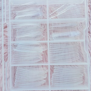 May include: A clear plastic storage box with multiple compartments filled with clear plastic nail tips.