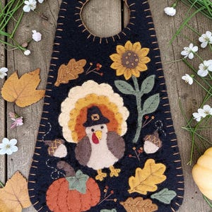 May include: A black felt door hanger featuring a central turkey wearing a hat, surrounded by appliqued felt designs including a sunflower, a pumpkin, oak leaves, and acorns. The edges are finished with tan blanket stitching.