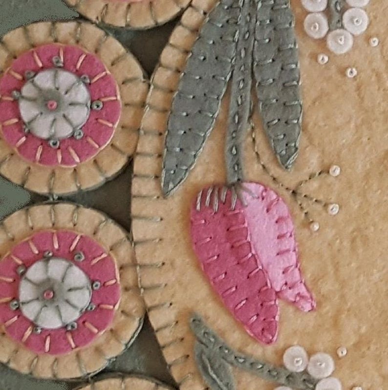 May include: A close-up of a handmade felt wall hanging with a pink flower, green leaves, and white beads. The background is a light beige felt.