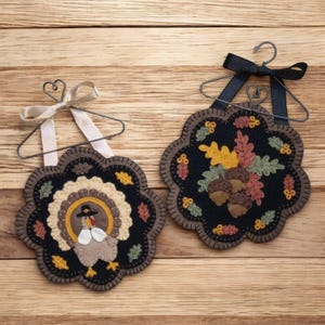 May include: Two felt wall hangings with a Thanksgiving theme. One features a turkey with a pilgrim hat and the other features a cluster of acorns and leaves.