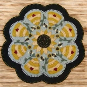 May include: A round, felted coaster with a floral design. The coaster features a central sunflower surrounded by yellow beehives, bees, and green leaves on a light blue background. The edge is black.