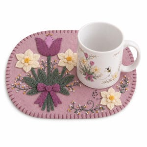 May include: A white ceramic mug with a floral design and a bee, placed on a pink felt placemat. The placemat features embroidered flowers and a purple bow. The mug and placemat create a charming, coordinated set.