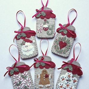 May include: Handmade Christmas ornaments in a jar shape. Each ornament is gray with a red scalloped top and a red ribbon. Designs include a Santa, a gingerbread man, candy canes, snowmen, a heart wreath, and the word "Joy".