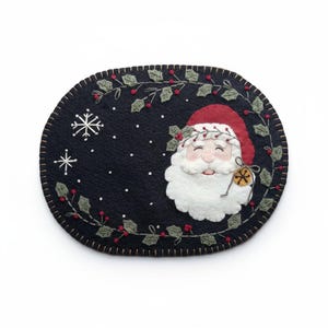 May include: An oval, black felt Christmas placemat featuring a smiling Santa Claus with a white beard and red hat. The placemat is decorated with embroidered snowflakes, holly, and berries. A wooden key hangs from Santa's beard.