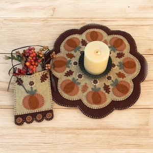 May include: A set of two felt autumn-themed table decorations. The larger piece is a round, scalloped mat with a brown border and a beige center featuring pumpkins, sunflowers, and leaves. The smaller piece is a beige pocket with a brown border and a pumpkin design. A white candle sits in the center of the mat.