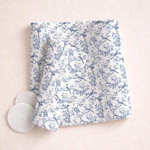 May include: White fabric with a blue floral and bird pattern. The fabric is folded and has a soft texture. Three white, round cotton pads are next to the fabric. The overall theme is home decor.