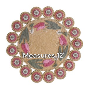 May include: A round felt rug with a floral design featuring pink tulips and green leaves. The rug is surrounded by a border of 12 smaller felt circles with white and pink accents. The rug measures 12 inches in diameter. "Measures 12" is written in black text on the rug.