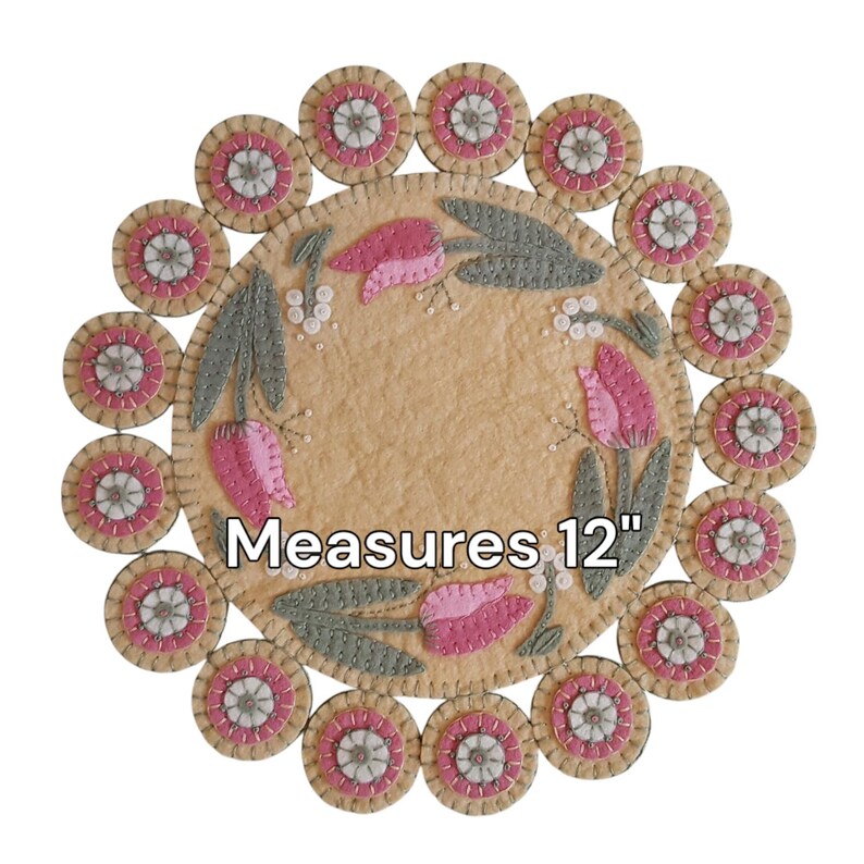 May include: A round felt rug with a floral design featuring pink tulips and green leaves. The rug is surrounded by a border of 12 smaller felt circles with white and pink accents. The rug measures 12 inches in diameter. "Measures 12" is written in black text on the rug.