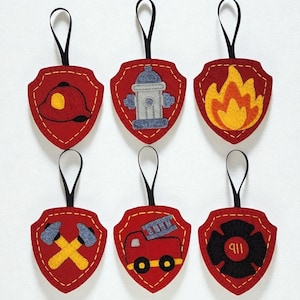May include: Six red felt firefighter shield ornaments with black ribbon hangers. Each shield features a different design: a helmet, a fire hydrant, flames, crossed axes, a fire truck, and a 911 emblem. The ornaments are stitched with yellow thread.