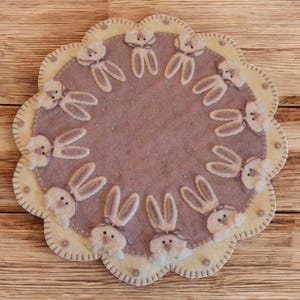 May include: A round, cream-colored felt table runner with a scalloped edge and a circle of white felt bunnies with pink ears around the center. The center of the runner is a light purple felt with a dotted pattern.