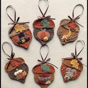 May include: Six felt acorn ornaments with brown ribbon hangers. Each acorn features autumn-themed designs, including leaves, pumpkins, and a squirrel, in shades of orange, brown, and yellow. These handmade ornaments are perfect for fall decor.