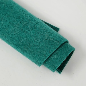 May include: A rolled-up piece of teal felt fabric. The felt has a textured, fibrous surface and is folded over on itself, revealing multiple layers. The color is a deep, saturated teal, and the fabric appears soft and pliable.