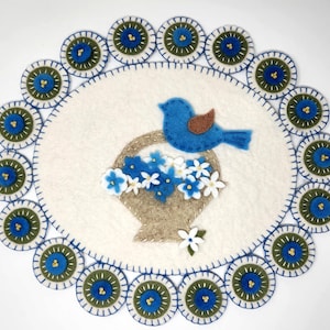 May include: A blue bird carrying a basket of white and blue flowers sits on a white felt circle surrounded by a border of green and blue circles.
