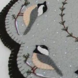 May include: A gray felt needlepoint wall hanging with a scalloped edge. The design features three black-capped chickadees perched on branches with a snowy background.