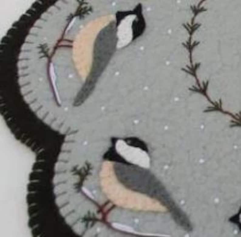 May include: A gray felt needlepoint wall hanging with a scalloped edge. The design features three black-capped chickadees perched on branches with a snowy background.