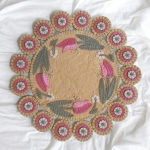 May include: A round wool rug with a beige center and a border of pink and white circles. The center features a design of pink tulips with green leaves and white beads.