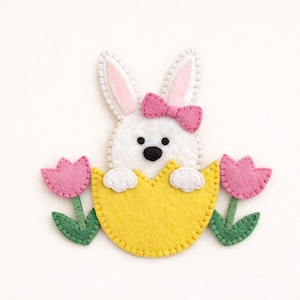 May include: A white felt bunny with pink bow and ears peeks out of a yellow egg. Two pink felt tulips with green stems and leaves flank the bunny, creating a spring or Easter theme.