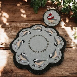 May include: A gray felt round table runner with a black border and a wreath of green leaves in the center. Eight small gray birds with yellow breasts are perched on branches around the wreath. A smaller gray felt ornament with a single bird wearing a red Santa hat is also included.