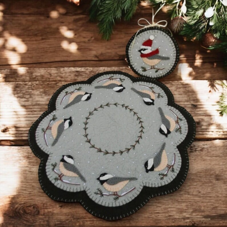 May include: A gray felt round table runner with a black border and a wreath of green leaves in the center. Eight small gray birds with yellow breasts are perched on branches around the wreath. A smaller gray felt ornament with a single bird wearing a red Santa hat is also included.