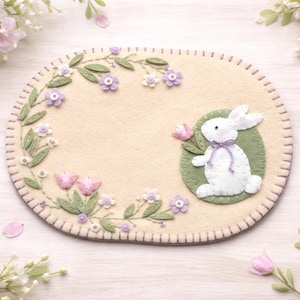 May include: An oval-shaped beige felt placemat with a stitched border. It features a white bunny with a purple bow, surrounded by green leaves, purple and white flowers, and pink butterflies. Decorative Easter theme.