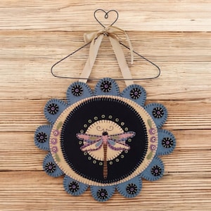 May include: A round felt wall hanging with a blue and black floral design. The center of the hanging features a yellow and pink dragonfly with brown legs.