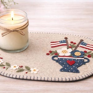 May include: A lit candle in a glass jar with a twine bow sits on a felt oval mat. The mat features a floral design and a patriotic motif with American flags and a blue vase with a heart.