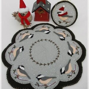 May include: A gray felt Christmas ornament with a black border and a design of eight birds wearing Santa hats. The birds are perched on branches with snow on the ground. The ornament is surrounded by a wreath of green branches.