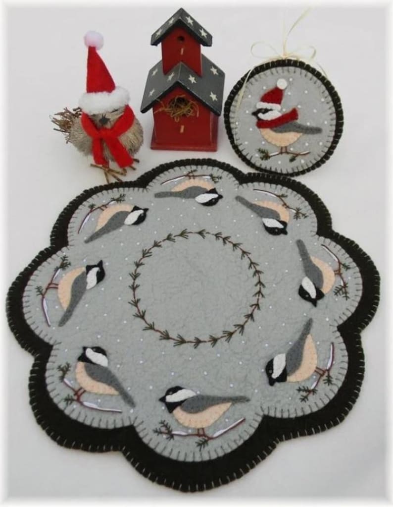 May include: A gray felt Christmas ornament with a black border and a design of eight birds wearing Santa hats. The birds are perched on branches with snow on the ground. The ornament is surrounded by a wreath of green branches.